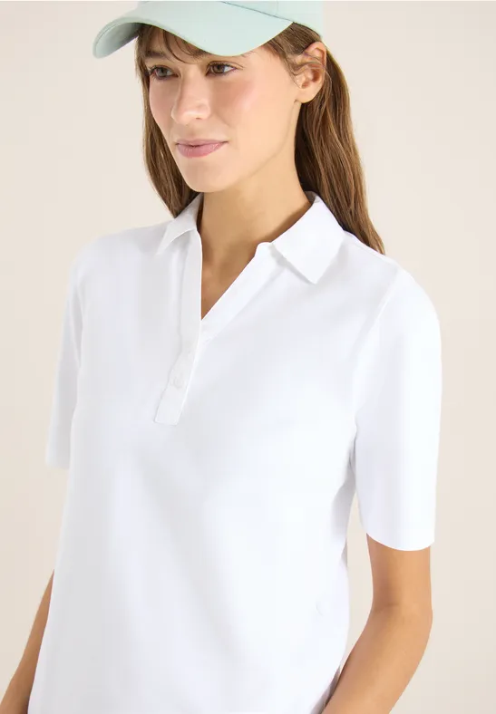 Eine Frau trägt ein weißes Polo-Shirt, hat einen lockeren Pferdeschwanz und steht vor hellem Hintergrund.