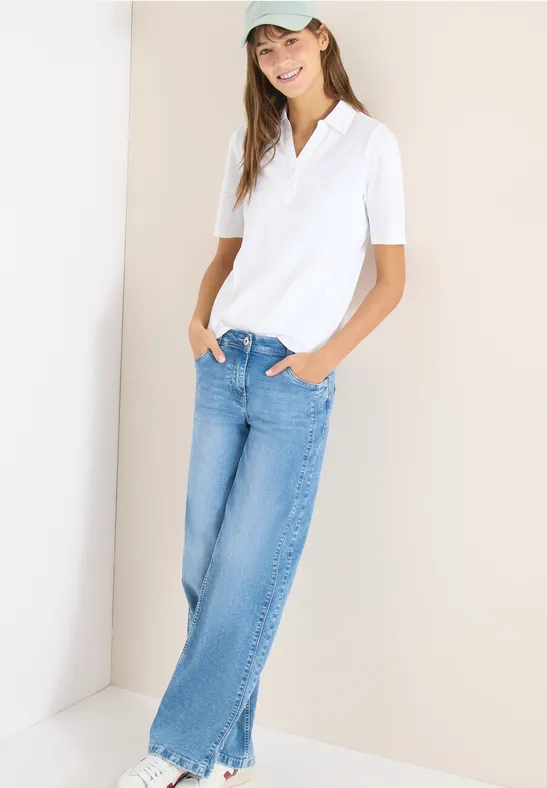 Eine Frau trägt weiße Polo-Shirt, blaue Jeans und Cap, steht entspannt mit Händen in den Taschen an einer Wand.