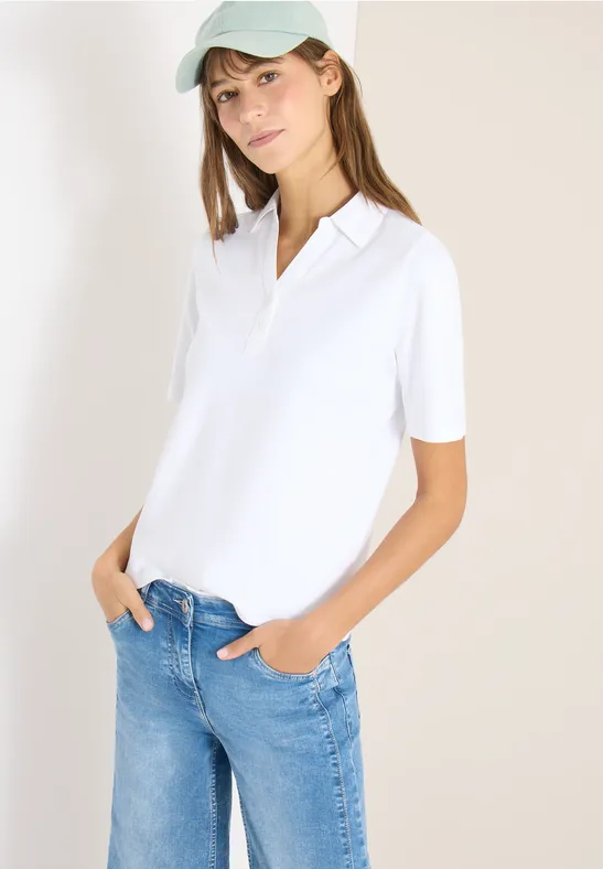 Frau trägt weißes Poloshirt und blaue Jeans, steht entspannt mit Händen in den Taschen