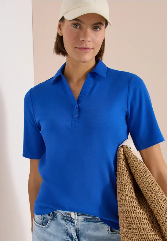Frau in knallblauem Poloshirt und hellen Jeans, hält beiges Strickjäckchen lässig über den Arm.