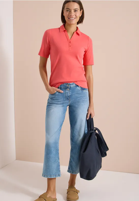 Frau trägt korallefarbenes Poloshirt, hellblaue Cropped-Jeans und Loafers, sie hält eine dunkelblaue Tasche.