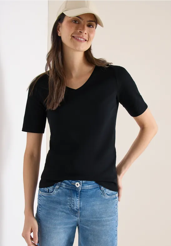 Frau in schwarzem T-Shirt und Jeans steht vor hellem Hintergrund, eine Hand in der Hüfte