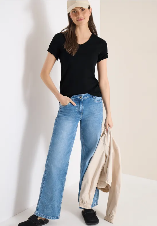 Modell trägt schwarzes T-Shirt, helle Jeans und hält eine beige Jacke, steht vor hellem Hintergrund.
