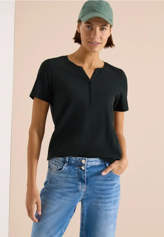 Frau trägt schwarzes T-Shirt und blaue Jeans, lässige Pose mit einer Hand in der Hosentasche.