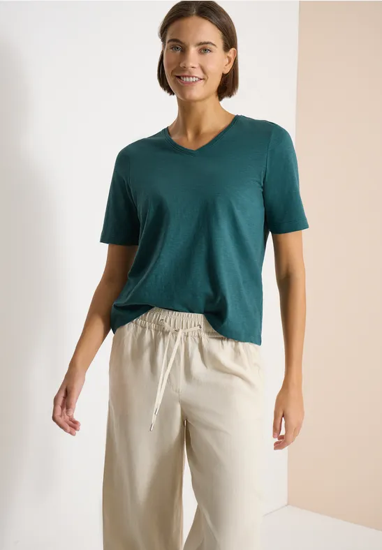 Frau in dunkelgrünem V-Shirt und beigen, weiten Stoffhosen vor neutralem Hintergrund.