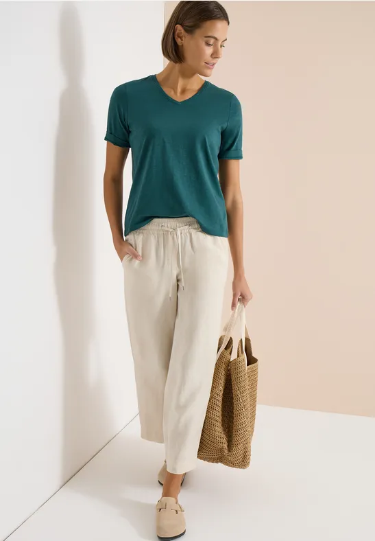 Frau in türkisfarbenem T-Shirt, weißer Leinenhose und Korbtasche.