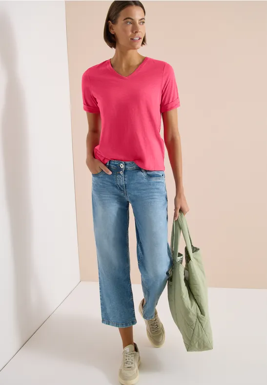 Model trägt pinkes V-Neck-Shirt, weite blaue Jeans und weiße Sneaker, hält grüne Jacke in der Hand.