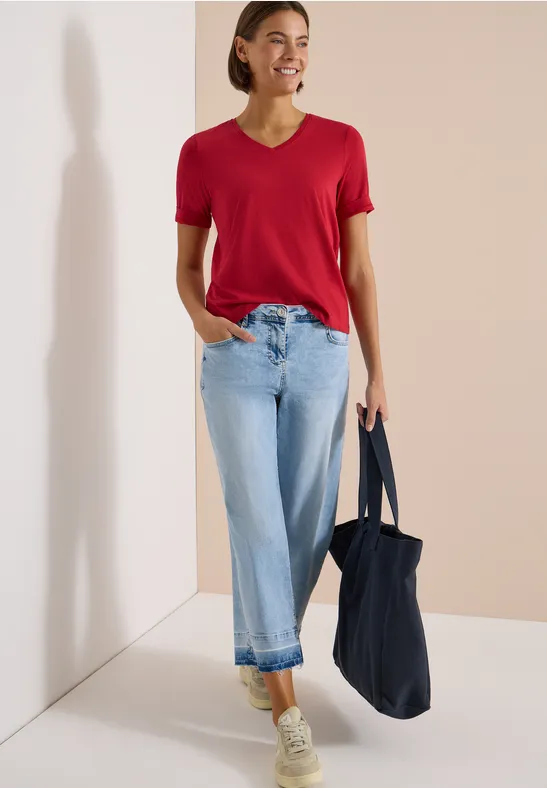 Eine Person mit rotem T-Shirt, hellblauen Jeans, weißen Sneakern und dunkelblauer Tasche vor hellem Hintergrund.