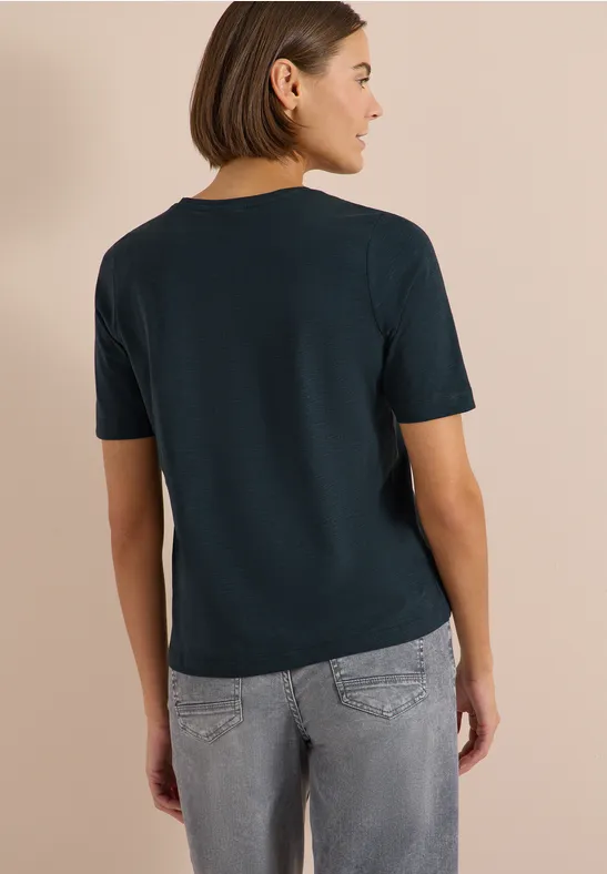 Frau von hinten in marineblauem T-Shirt und grauer Jeans vor beigem Hintergrund.