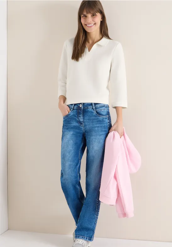 Person in weißem Pullover und Jeans hält rosa Jacke vor neutralem Hintergrund.