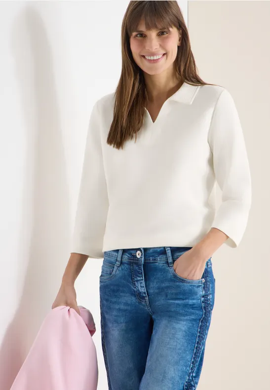 Frau vor weißer Wand in weißem Pullover, blauen Jeans und rosa Jacke in der Hand.