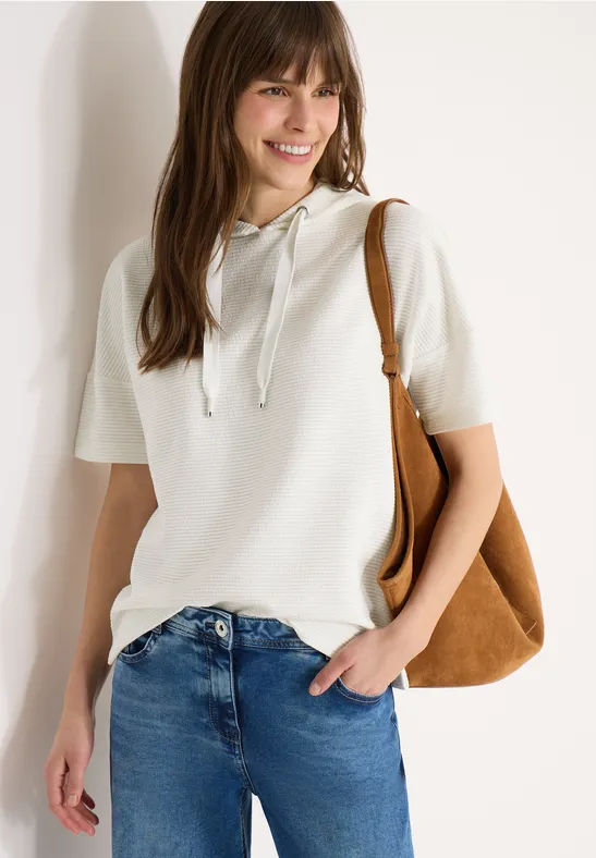 Eine Frau lehnt an einer weißen Wand, trägt weißen Hoodie, blaue Jeans und braune Schultertasche.