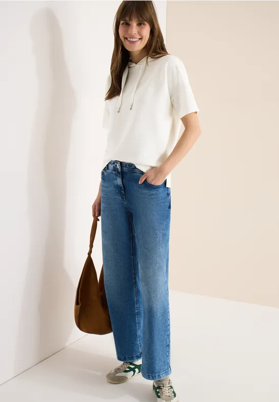Eine Person steht vor heller Wand, trägt weißes T-Shirt, weite blaue Jeans und hält eine braune Tasche.