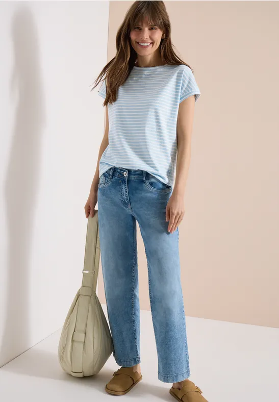 Frau in hellblau-weiß gestreiftem Shirt, gerader Jeans und großer, lässiger Tasche vor neutralem Hintergrund.