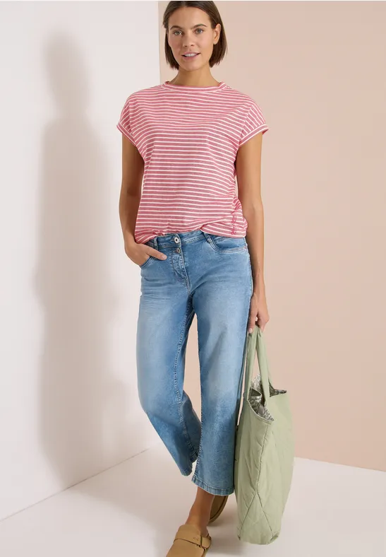Eine Frau trägt eine rote gestreifte Bluse, weite blaue Jeans und hält eine grüne Tasche.