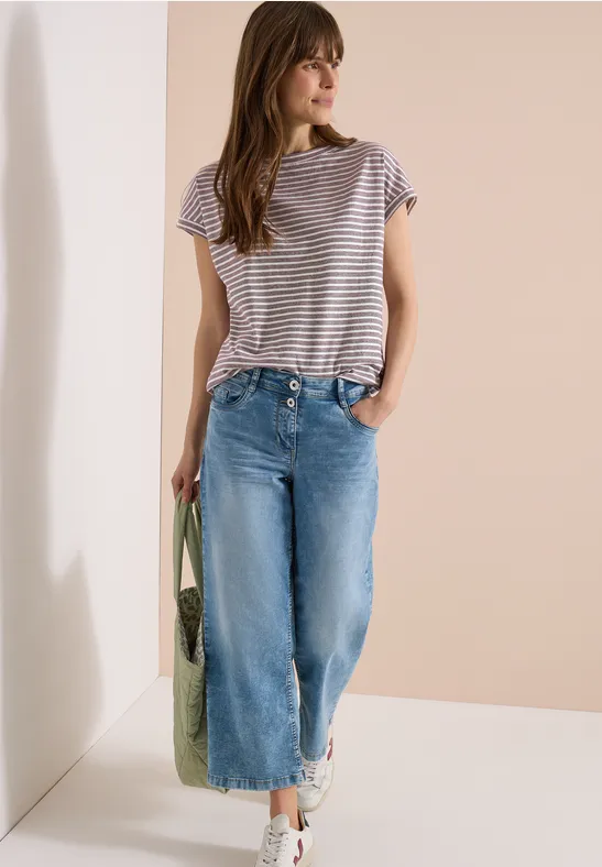 Frau trägt gestreiftes Shirt, weite Jeanshose, weiße Sneaker und grünen Netzbeutel vor beiger Wand.