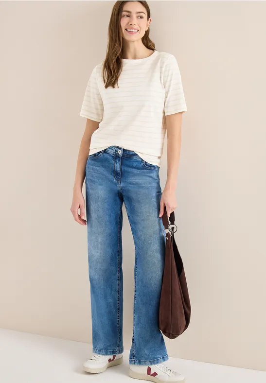 Frau trägt weißes gestreiftes T-Shirt, weite blaue Jeans und eine Tasche vor neutralem Hintergrund.
