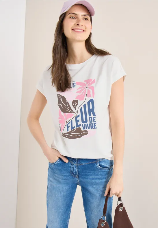 Frau trägt weißes T-Shirt mit Blumenprint 'FLEUR DE VIVRE', blaue Jeans und Handtasche.