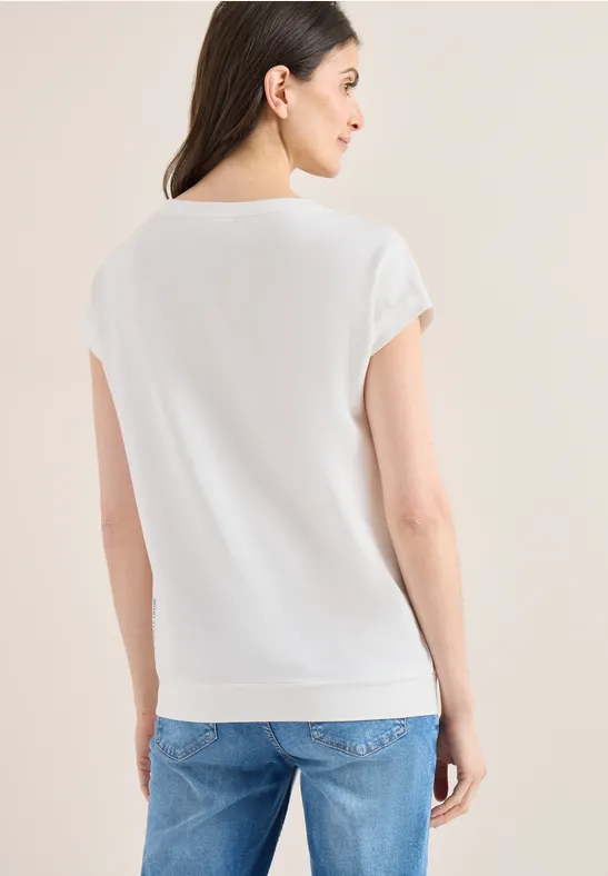 Frau von hinten in weißem T-Shirt mit umgeschlagenen Ärmeln und heller Jeans vor hellem Hintergrund.
