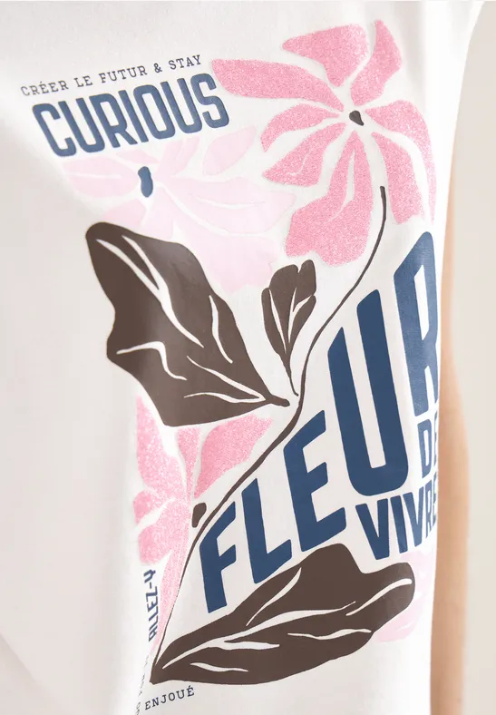 Nahe Ansicht eines T-Shirts mit rosa Blume, braunen Blättern und blauem Text 'Curious' und 'Fleur Vive'.