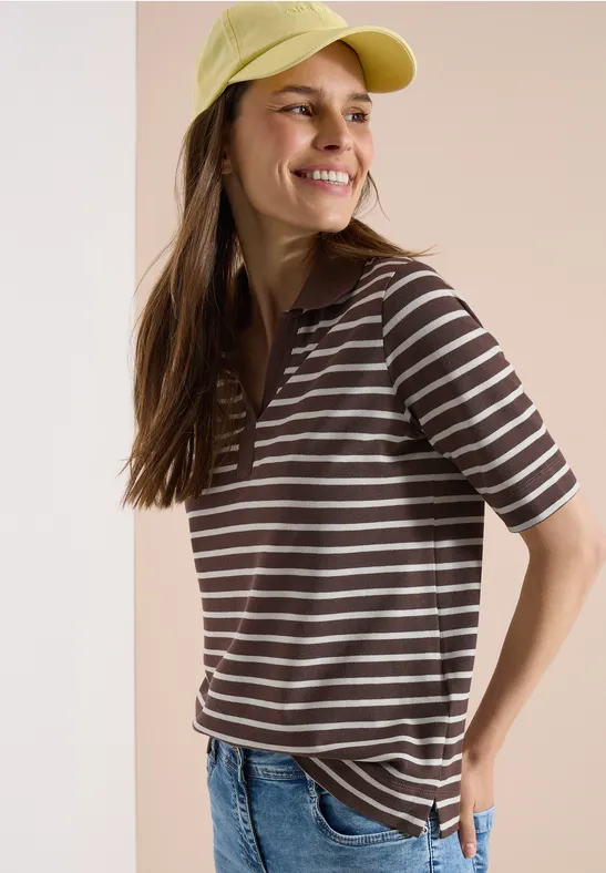 Une personne porte une casquette jaune, un t-shirt rayé marron et blanc et un jean, sur un fond neutre.