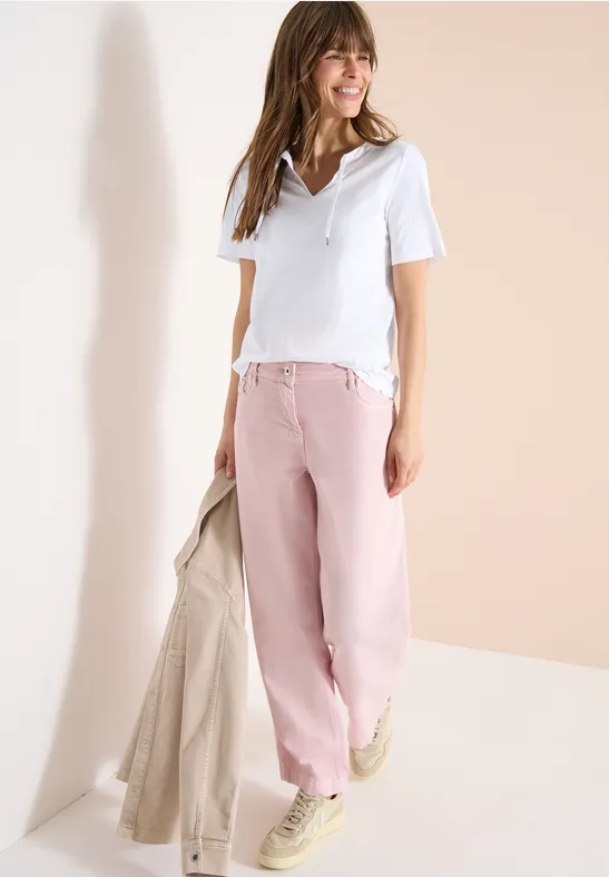 Eine Frau in weißem T-Shirt, rosa weiter Hose, hellen Sneakern, hält eine beige Jacke.