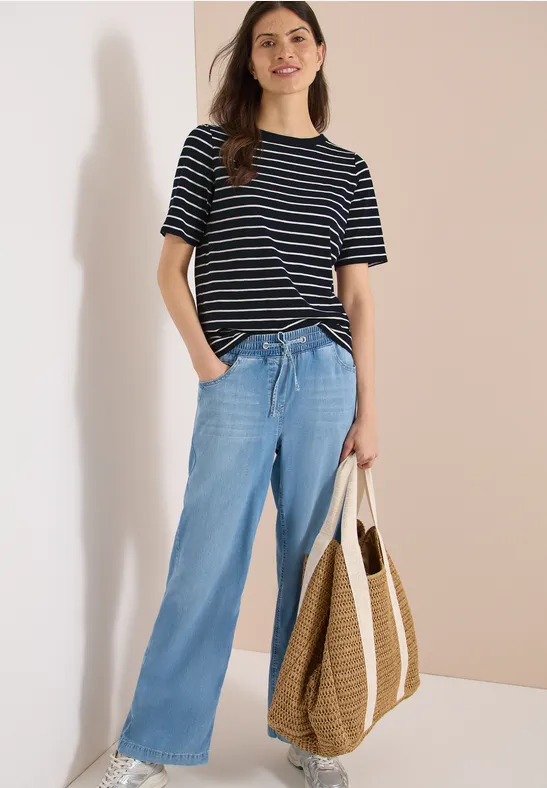 Eine Person trägt ein gestreiftes T-Shirt, weite Jeans und hält eine geflochtene Tasche.