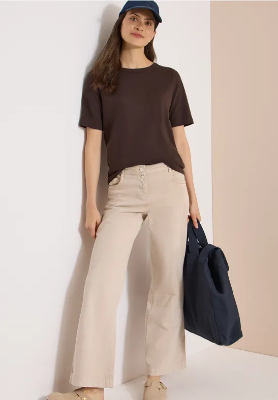 Frau lehnt an einer Wand, trägt dunkelbraunes T-Shirt, beige, weite Hose und hält eine dunkelblaue Tasche.