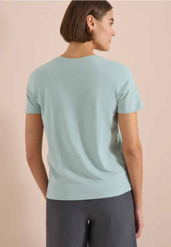 Frau von hinten in hellblauem T-Shirt und dunkler Hose vor neutralem Hintergrund.