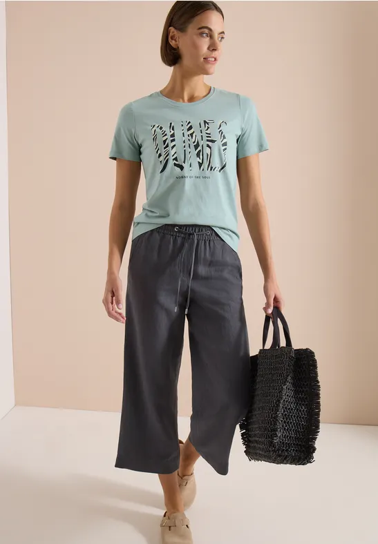 Frau trägt mintgrünes T-Shirt mit Schriftzug, weit geschnittene dunkle Culotte, Espadrilles und schwarzen Tote-Bag.