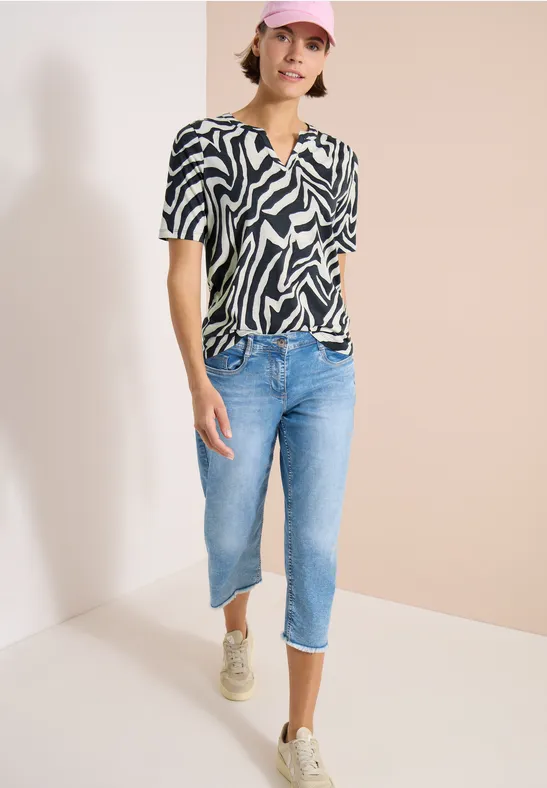 Person trägt schwarz-weißes Zebra-Muster-Shirt, blaue Cropped-Jeans und Sneaker vor neutralem Hintergrund.