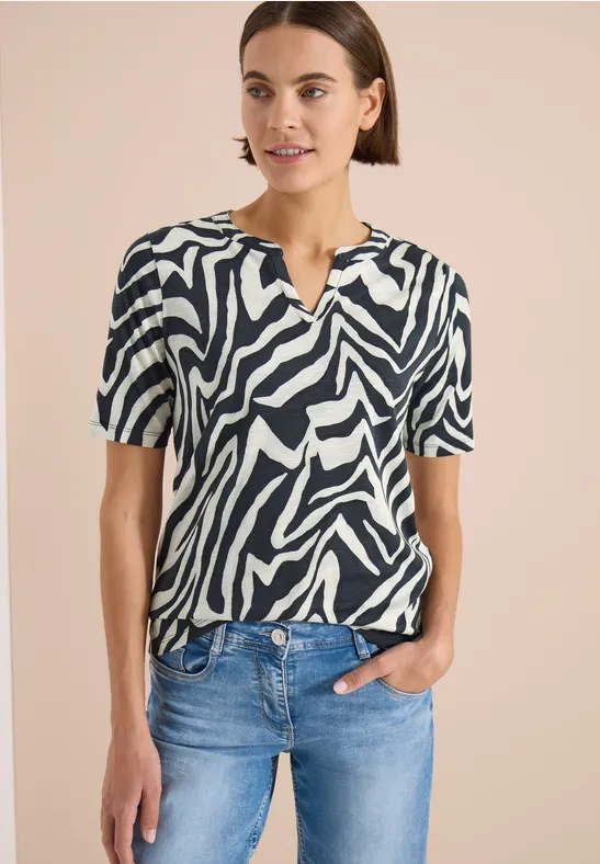 Frau trägt kurzärmliges, schwarz-weißes Zebramuster-Shirt und blaue Jeans, Hand in Tasche.