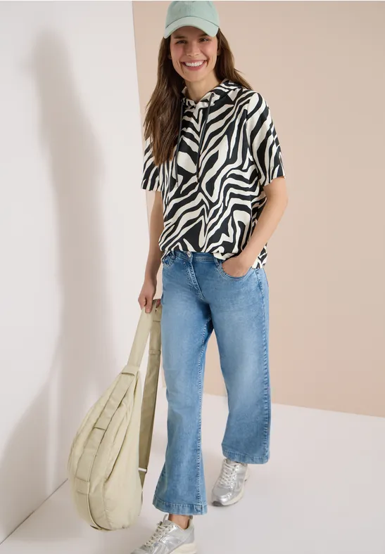 Eine Frau in Zebra-Shirt, weiten Jeans, silbernen Sneakers und großer Tasche vor hellem Hintergrund.