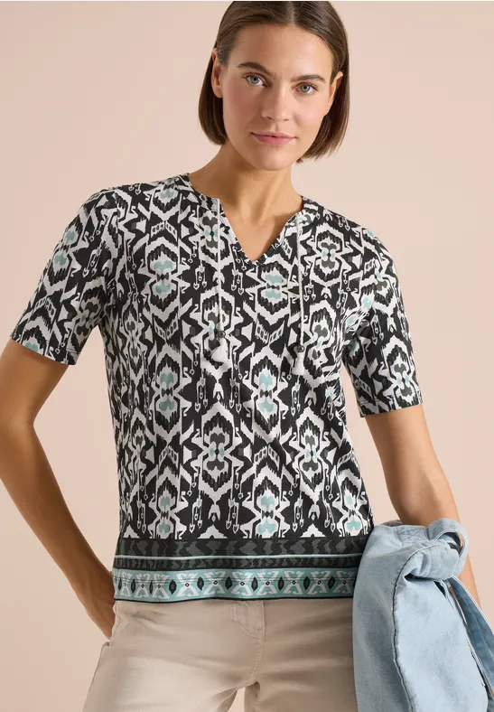 Damenbluse mit kurzem Ärmel, geometrischem Schwarz-Weiß-Türkis-Muster und V-Ausschnitt, dazu leichte Jeansjacke.