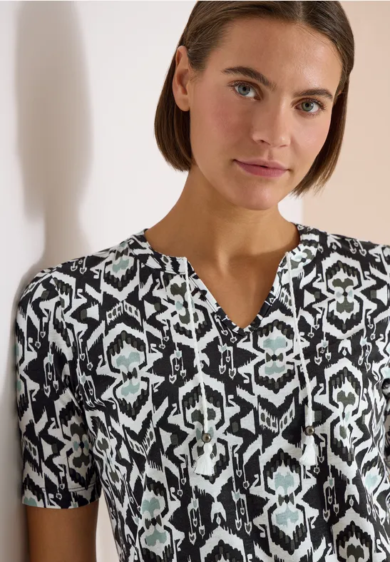 Frau in schwarz-weißer, kurzärmliger Bluse mit geometrischem Muster und dezentem V-Ausschnitt.