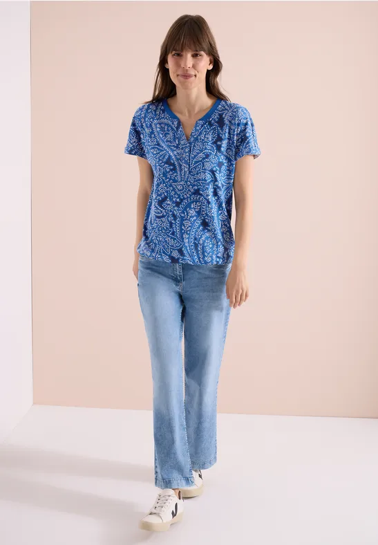 Femme en chemisier bleu à motifs, jean bleu et baskets blanches devant un fond beige.