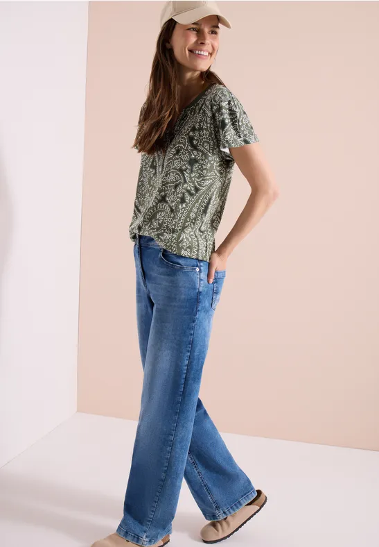 Frau in grüner, gemusterter Bluse, blauen Jeans und braunen Schuhen steht seitlich vor hellem Hintergrund.