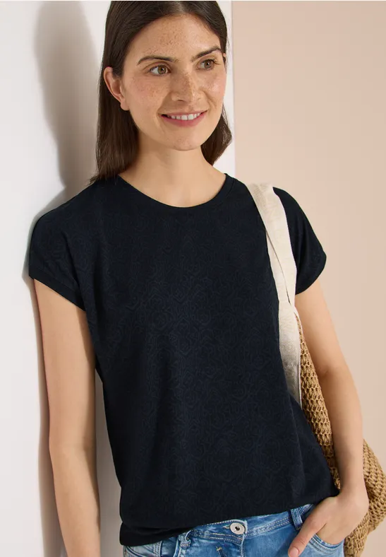 Person lehnt an Wand, trägt schwarzes T-Shirt und Strohschultertasche.