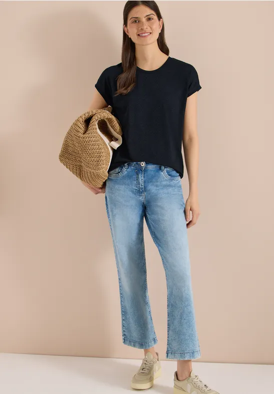 Frau vor beiger Wand in schwarzem T-Shirt, hellen Jeans, Sneakers und mit geflochtenem Strohkörbchen.