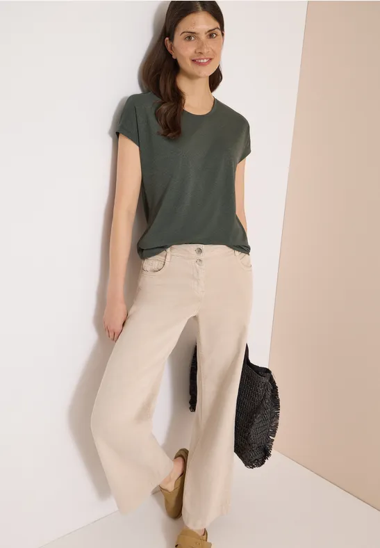 Eine Frau lehnt an weißer Wand, trägt dunkelgrünes T-Shirt, helle weite Jeans, goldene Schuhe und schwarze Tasche.