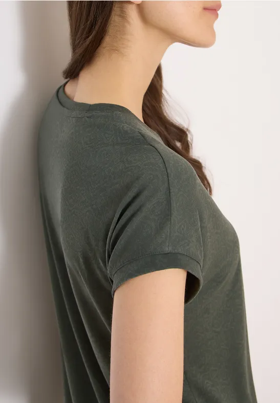 Junge Frau seitlich, lehnt an weißer Wand, trägt dunkelgrünes kurzärmeliges T-Shirt, Blick nach vorn