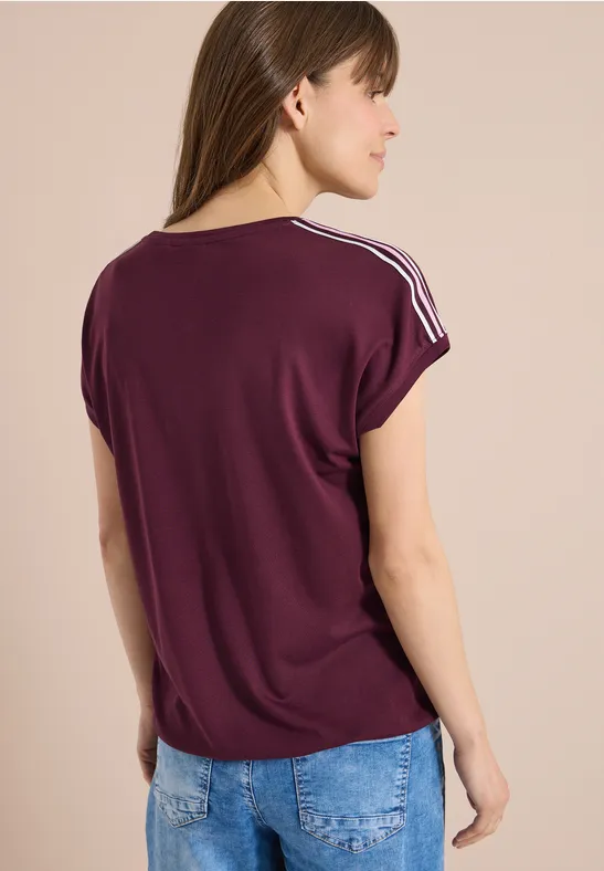 Frau von hinten in lockerem violettem T-Shirt mit weißen Streifen an den Schultern und heller Jeans.