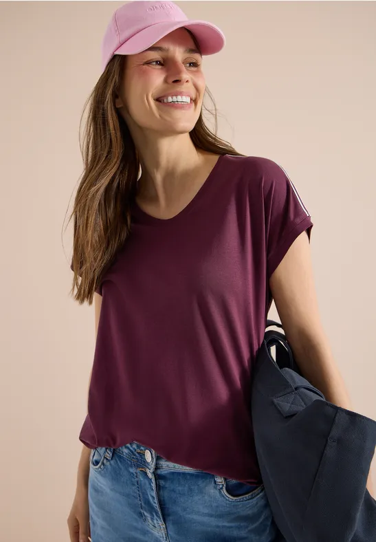 Frau mit lila T-Shirt, Jeans, rosa Cap, trägt blaue Tasche vor beigem Hintergrund.