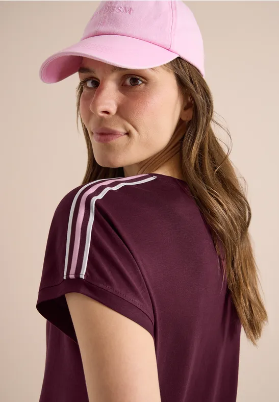 Eine Person von hinten trägt ein bordeauxrotes T-Shirt mit weißen Schulterstreifen und eine rosa Baseballkappe.