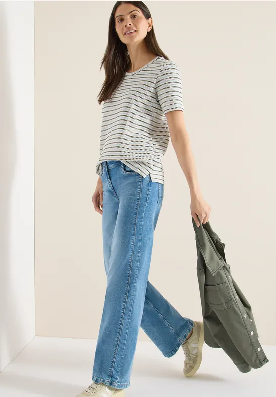 Frau in gestreiftem T-Shirt, weiter Jeans und Sneakern, hält eine grüne Jacke in der Hand.