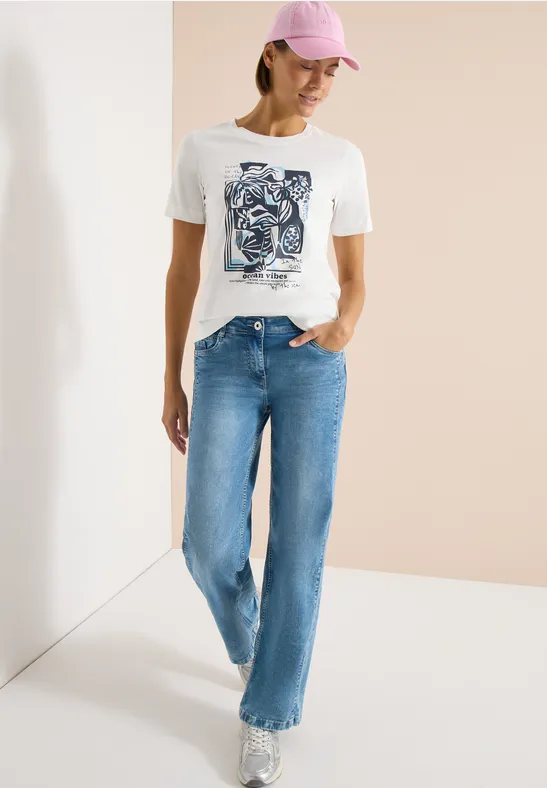 Frau in weißem Shirt mit Print und blauen Jeans lehnt lässig an einer Wand.