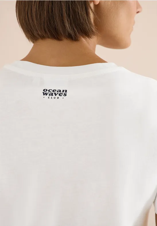 Rückenansicht einer Frau mit kurzem braunem Haar in weißem T-Shirt mit kleinem 'ocean waves'-Logo am Nacken.