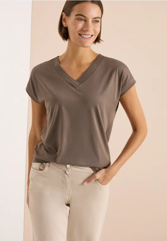 Frau trägt graubraunes V-Ausschnitt-T-Shirt und helle Hose, eine Hand in der Tasche.