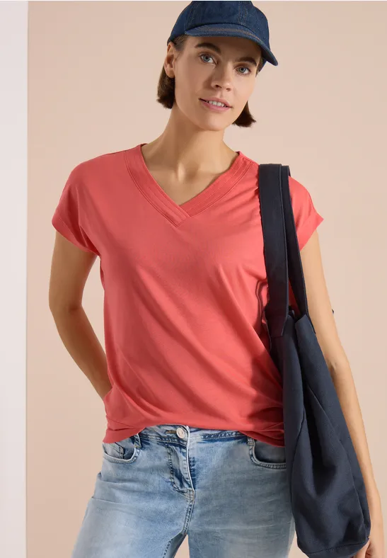 Frau in rotem V-Shirt, Jeans, dunkelblauer Stofftasche und Mütze vor beigem Hintergrund