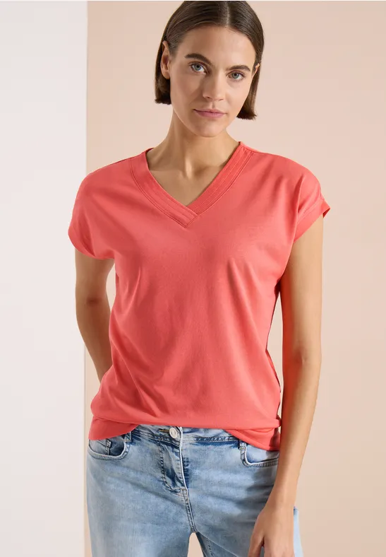 Frau trägt korallenfarbenes V-Shirt und Jeans vor zweifarbigem Hintergrund.